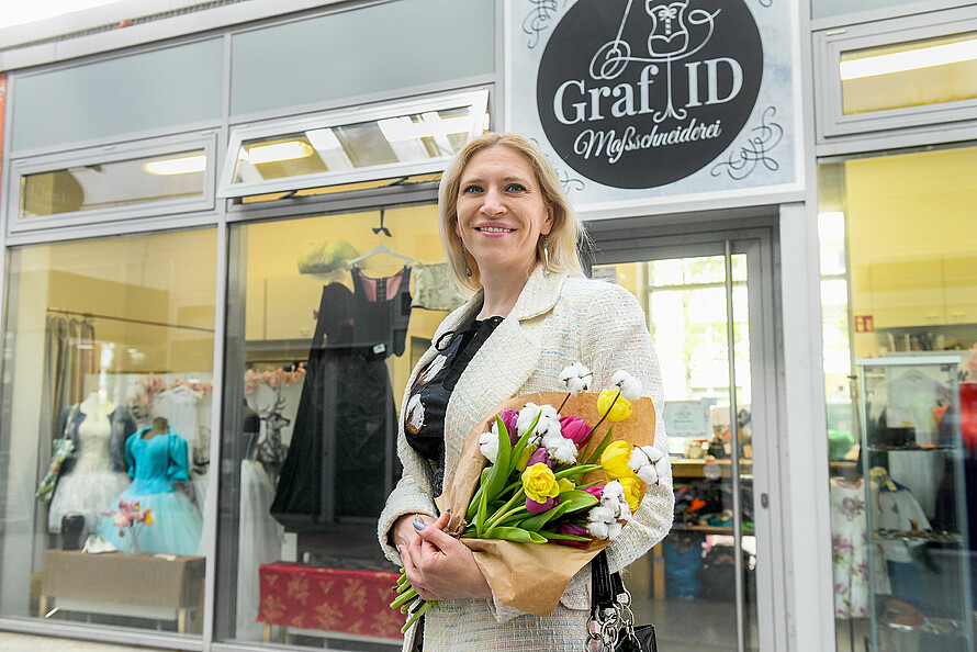 Die Schneiderin Darya Graf steht vor ihrem Atelier auf der Marzahner Promenade. Sie hält einen Blumenstrauss in den Armen und lächelt in die Kamera. Im Schaufenster des Ateliers sind Kostüme zu erkennen.