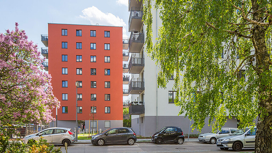 Ansicht eines rotfarbigen Neubaus in der Walter-Franck-Zeile in der Berliner Gropiusstadt. Im Vordergrund ist eine Straße, an der Autos geparkt sind sowie zwei Bäume, die blühen und Blätter tragen.