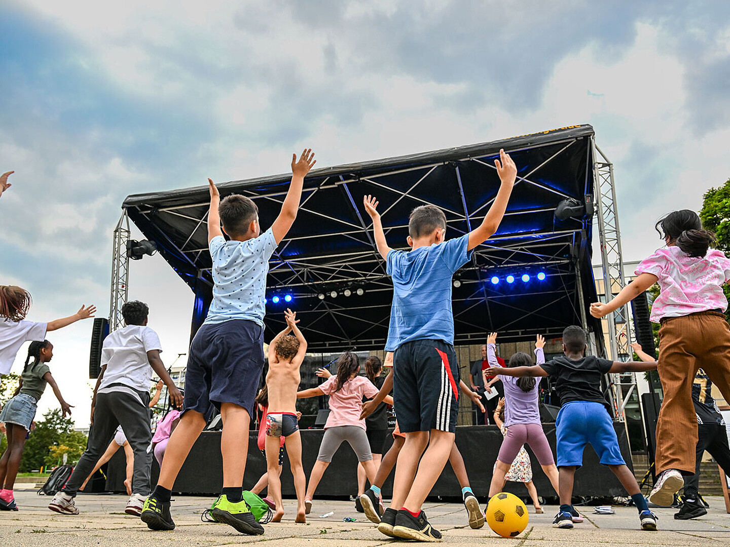 Kinder stehen vor einer Bühne vor freiem Himmel und machen Sport bzw. Tanzen zum Programm.