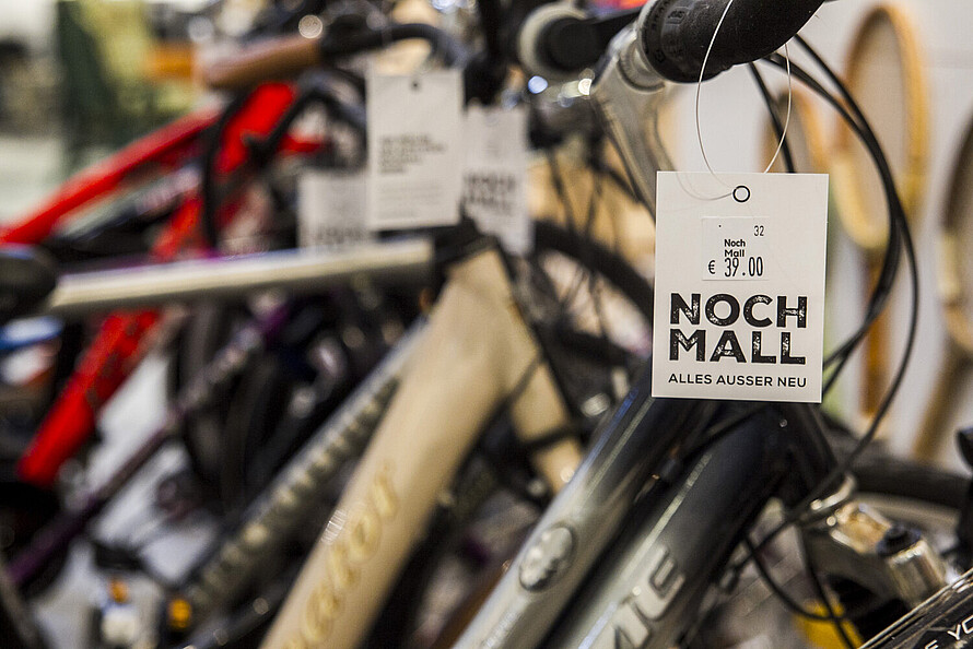 Auf dem Bild sind mehrere Fahrräder zu sehen, die in einem Geschäft zum Verkauf angeboten werden. Im Vordergrund hängt ein Preisschild an einem Fahrradlenker, auf dem "Noch Mall" und "Alles außer neu" steht. Der Preis beträgt 39 Euro. Die Fahrräder im Hintergrund sind teilweise unscharf und das Schild steht im Fokus des Bildes.