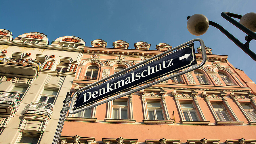 Im Hintergrund Gebäude mit Stuck und Verzierungen, im Vordergrund ein Schild mit der Aufschrift „Denkmalschutz“.