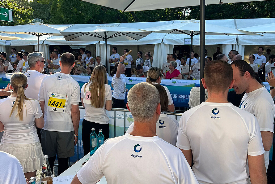 Das Foto zeigt Läufer bei einem Teamstaffellauf in Berlin. Sie stehen mit dem Rücken zur Kamera und haben ein Logo von degewo auf dem Laufshirt. Sie stehen hinter einer Bande und jubeln den einlaufenden Kolleginnen und Kollegen zu.