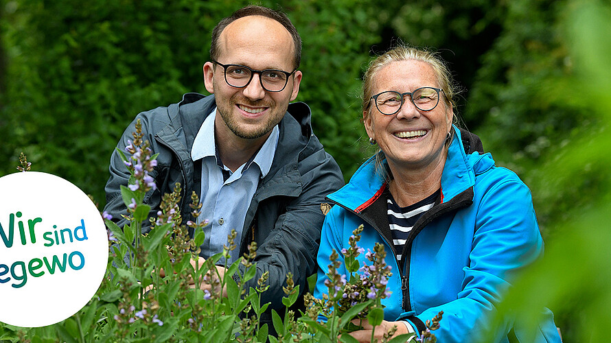 Zwei Mieter sitzen im hohen Gras ihres Gemeinschaftsgartens und lächeln in die Kamera.