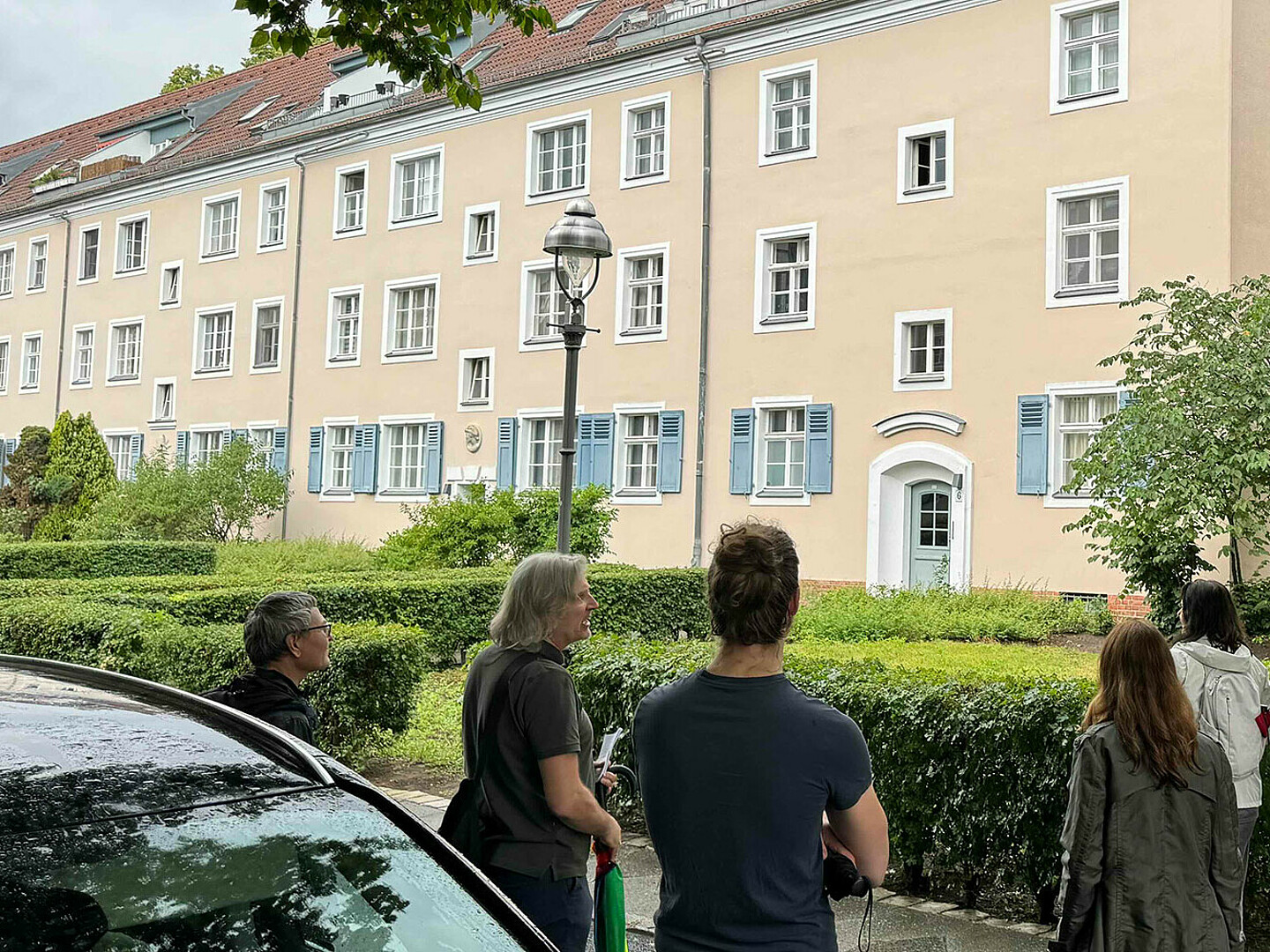 Personen nehmen an einer Führung zum Thema Stadtarchitektur teil. Sie stehen auf einer Straße und schauen sich das Gebäude gegenüber an. Dabei handelt es sich um den denkmalgeschützten Schillerhof in Berlin.