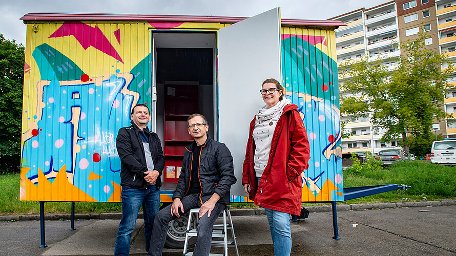 Drei Personen stehen vor einem bemalten Container.