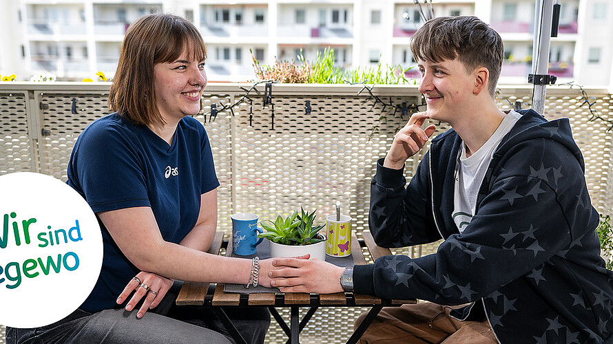 Ein junges Paar sitzt auf dem Balkon auf Stühlen gemeinsam an einem Tisch. Sie schauen sich in die Augen und halten Händchen. Im Hintergrund sind verschwommen Gebäude im Plattenbaustil zu erkennen. Es ist Frühling, einige Pflanzen sind auf dem Balkon gepflanzt.