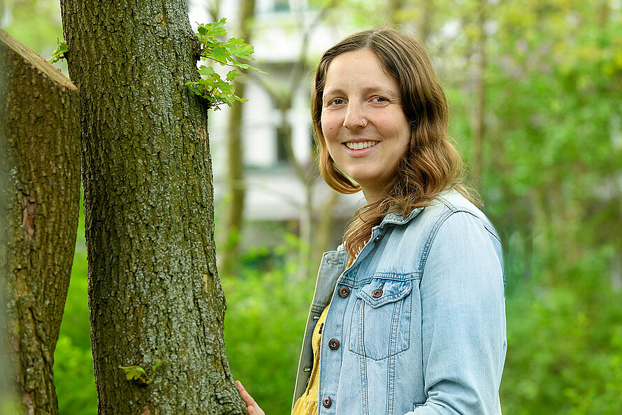 Eine Frau stütz sich mit den Händen an einen Baum ab und lächelt in die Kamera. 