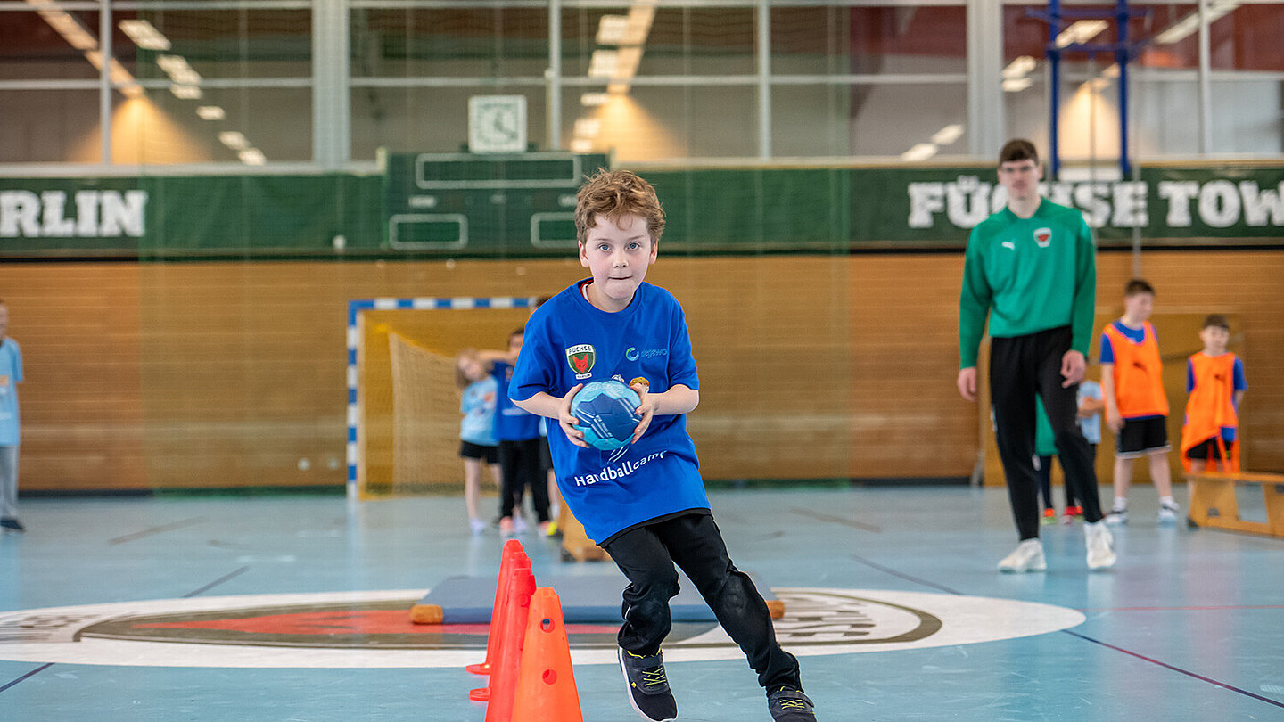 Ein Junge läuft mit einem Handball in der Hand um einen Parcours aus Hütchen. im Hintergrund beobachtet der Trainer. 