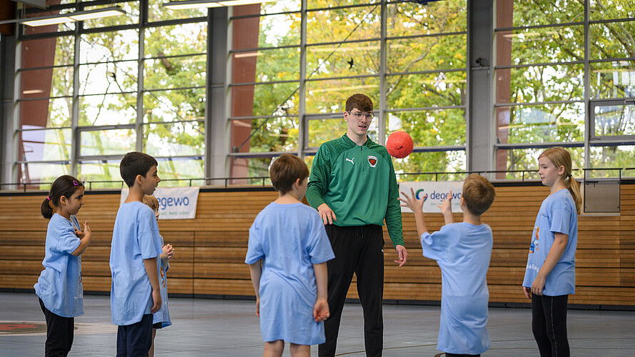 Eine Gruppe Kinder steht in einer Sporthalle mit ihrem Trainer. Ein Kind wirft einen kleinen roten Ball in Richtung Trainer.