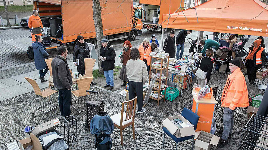 Das Bild zeigt eine Szene von einem BSR-Kieztag. Im Vordergrund stehen diverse Möbelstücke und Haushaltsgegenstände auf einem gepflasterten Platz. Mehrere Personen durchstöbern die Gegenstände oder unterhalten sich. Unter einem orangefarbenen Pavillon mit der Aufschrift “Berliner Stadtreinigung" und dem Logo der BSR sind weitere Gegenstände ausgestellt. Im Hintergrund sind orangefarbene BSR-Lastwagen und Mitarbeiter der BSR in orangefarbener Arbeitskleidung zu sehen. Die Atmosphäre wirkt geschäftig und gemeinschaftlich, während die Teilnehmenden die Gelegenheit nutzen, sich von nicht mehr benötigten Gegenständen zu trennen und diese weiterzuverwenden.