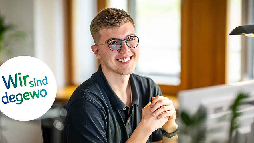 Ein junger Mann mit Brille sitzt am Schreibtisch und stützt seine Arme auf den Tisch. Er lächelt freundlich.