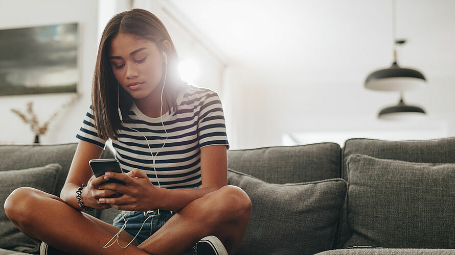 Ein Mädchen in schwarz-weiß Freizeitkleidung sitzt auf dem grauen Sofa und hört Musik über kabelgebundene In-Ear-Kopfhörer