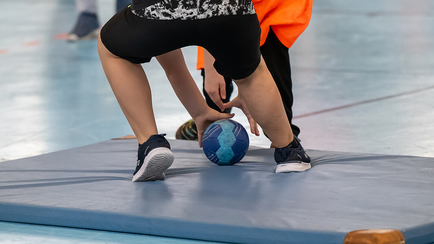 Eine Turnmatte liegt auf dem Boden einer Sporthalle. Zwei Kinder stehen darauf und versuchen, den Handball zu erlangen. 