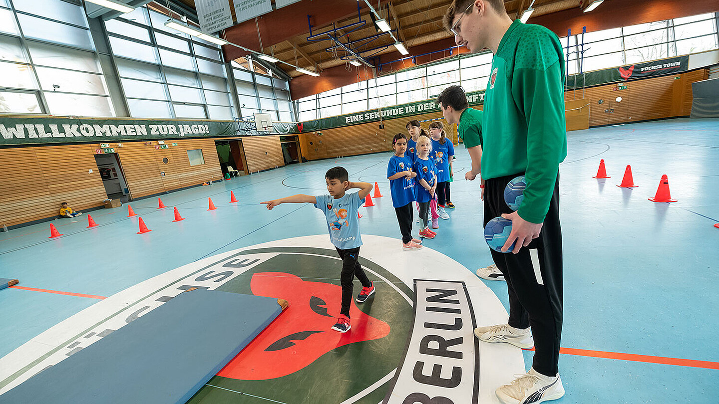 Ein Trainer übt mit einem Kind die richtige Technik, um den Handball auf das Tor zu werfen. 