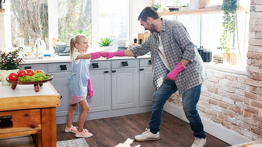 Das Foto zeigt Vater und Tochter mit pinken Gummihandschuhen in einer Küche. Beide lachen und machen einen Faustgruß.