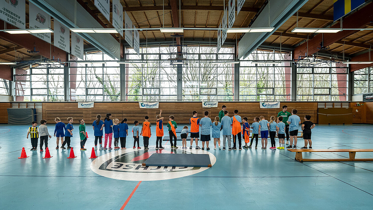 Viele Kinder stehen in einer Sporthalle in einer Reihe. Sie warten auf die nächste Anweisung des Trainers.