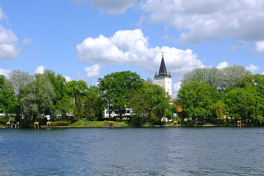 Zu sehen ist die Halbinsel Alt-Stralau in Berlin mit der darauf stehenden Dorfkirche. Der Blick wandert über das Wasser der Spree. Der Betrachter steht auf der Seite des Treptower Parks.