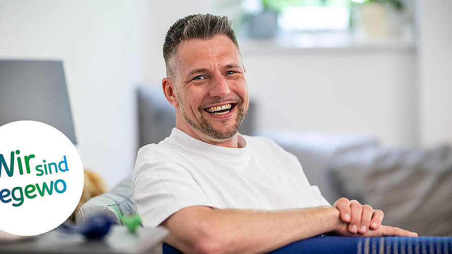 Mario Prymuschala sitzt in seiner Wohnung auf einer Couch. Seine Arme liegen auf der Lehne der Couch. Er lächelt in die Kamera. Im Hintergrund ist eine Stehlampe zu erkennen. 