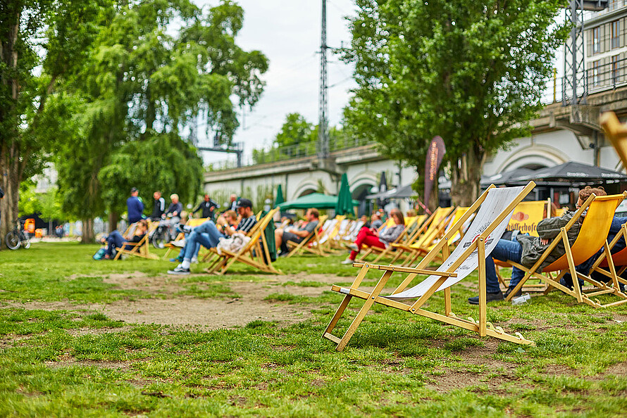 Liegestühle stehen auf der Wiese des James-Simon-Park in Berlin. Es ist Sommer. Vereinzelt sitzen Leute auf den Stühlen oder dem Rasen. Im Hintergrund der ist der S-Bahn-Bogen zu sehen.