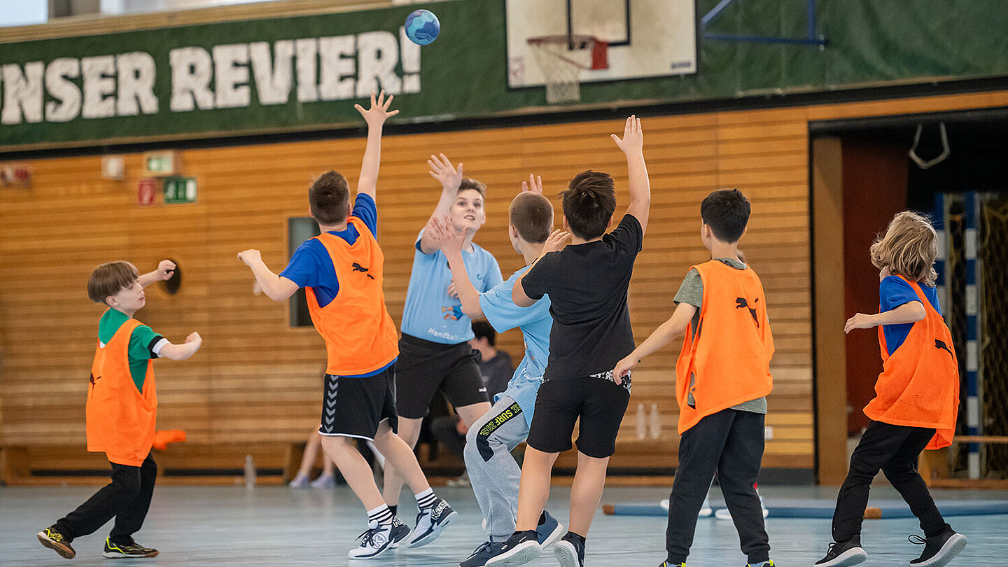 Eine Gruppe Kinder spielt in zwei Teams eingeteilt mit dem Handball in einer Sporthalle.