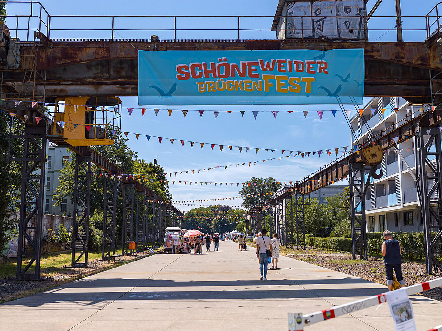 Zu sehen ist eine Straße in Berlin Schöneweide, auf der ein Straßenfest stattfindet. Über der Straße ist ein Banner gezogen mit dem Schriftzug "Schöneweider Brückenfest".