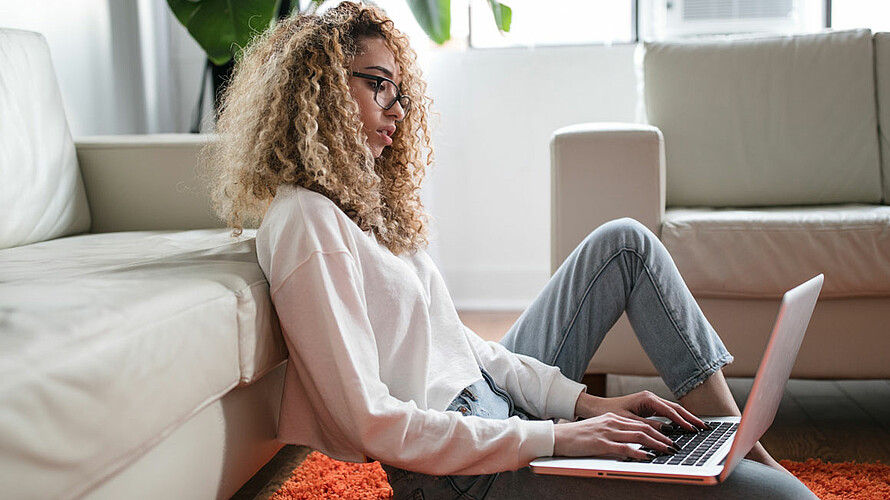 eine Frau, die sich an ein weißes Sofa lehnt und einen Laptop auf ihren Schoß legt.