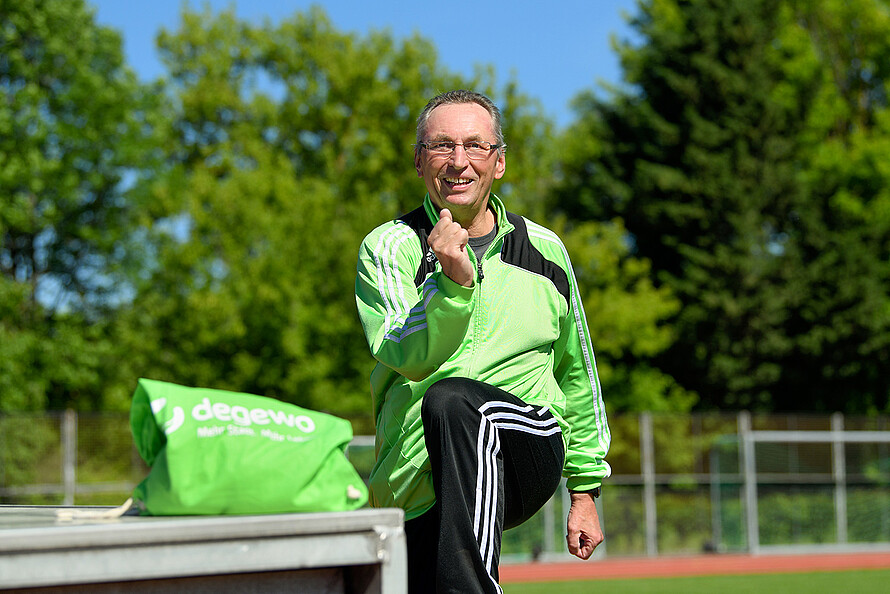 Das Bild zeigt Uwe Tolle bei der Aufwärmung auf einer Tartanbahn. Er trägt eine schwarze Trainingshose und eine grüne Laufjacke. Im Vordergrund ist ein Turnbeutel mit degewo-Logo zu sehen.