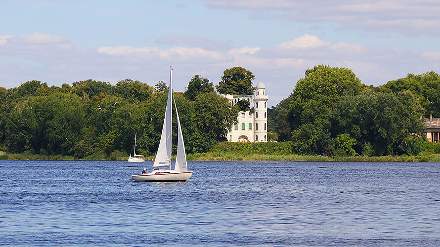 Zu sehen ist der Berliner Fluss Havel im Sommer. Mehrere Segelboote sind auf dem Wasser unterwegs. Im Hintergrund ist die Pfaueninsel und das darauf befindliche Schloss zu sehen. 