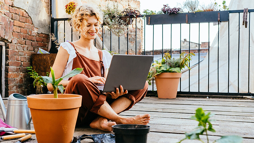 Eine blonde Frau sitzt auf einem Balkon im Schneidersitz und hat einen Laptop in der Hand. Um sie herum stehen Blumentöpfe und Werkzeuge zum Gärtnern.