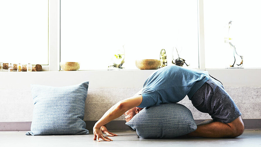 Ein Mann praktiziert Yoga in einer Backbend-Pose. er hat ein graues Kissen, um auf seinem Rücken zu ruhen. Hinter ihm befindet sich ein Fensterbrett, das mit einer Reihe von Schalen und Pflanzen geschmückt ist. Auf dem Boden liegt auch ein Kissen in der gleichen Farbe.