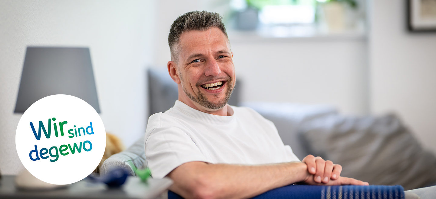 Mario Prymuschala sitzt in seiner Wohnung auf einer Couch. Seine Arme liegen auf der Lehne der Couch. Er lächelt in die Kamera. Im Hintergrund ist eine Stehlampe zu erkennen. 