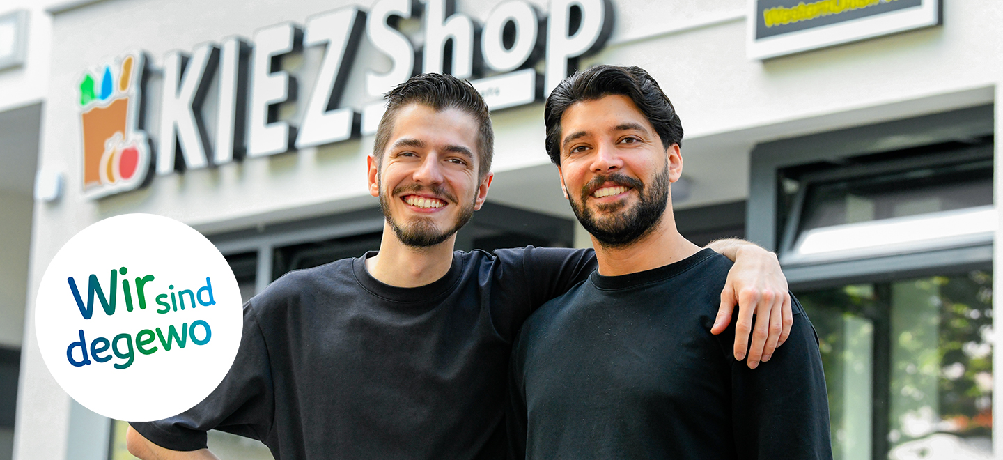 Zwei Männer stehen vor dem Eingang eines Kiezshops im Brunnenviertel in Berlin. Der eine Mann hat seinen Arm um die Schulter des anderen Mannes gelegt. Sie lächeln in die Kamera.