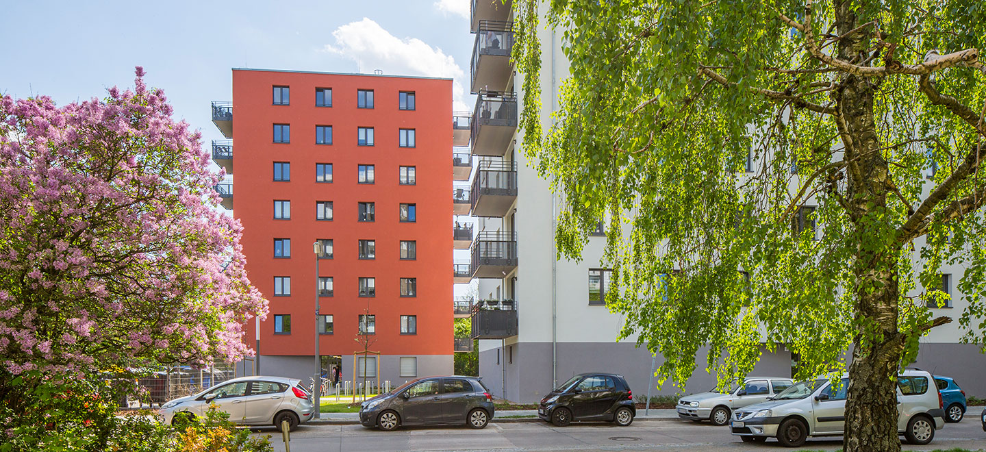 Ansicht eines rotfarbigen Neubaus in der Walter-Franck-Zeile in der Berliner Gropiusstadt. Im Vordergrund ist eine Straße, an der Autos geparkt sind sowie zwei Bäume, die blühen und Blätter tragen.